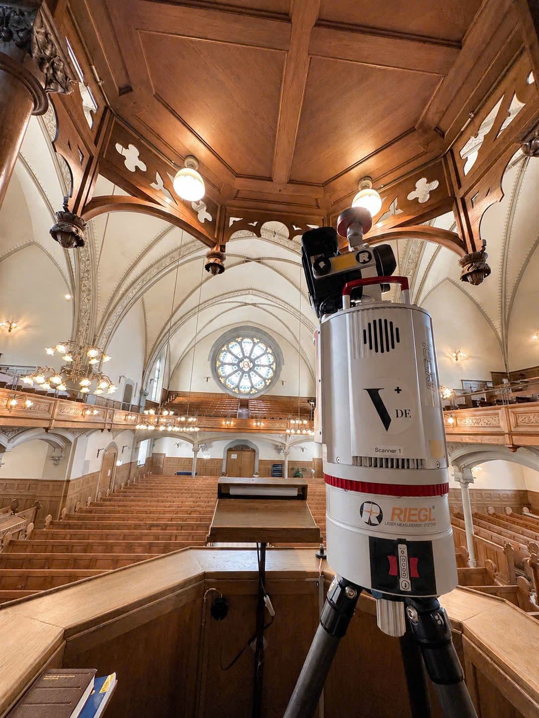 Innenansicht einer historischen Kirche mit Holzdecke, Emporen und Orgelraum; im Vordergrund steht ein Vermessungslaser/3D-Laserscanner auf Stativ, im Hintergrund Rundfenster und Kirchenbänke.