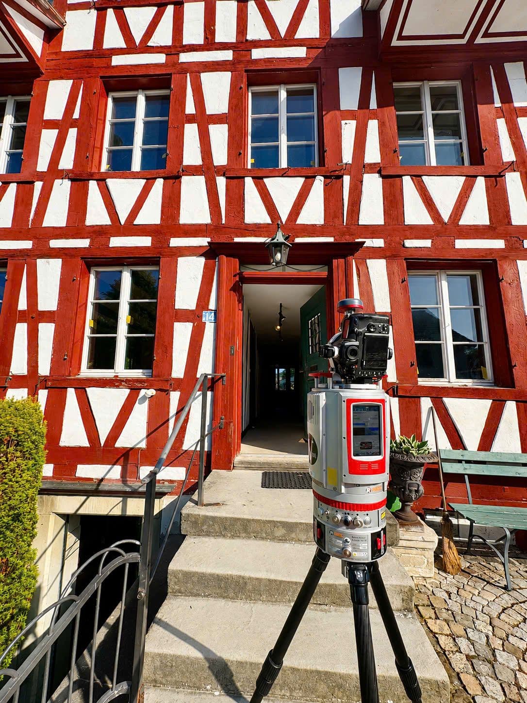 Rotes Fachwerkhaus mit weißem Putz und Eingangstreppe; davor steht ein 3D-Laserscanner auf Stativ zur Gebäudevermessung.