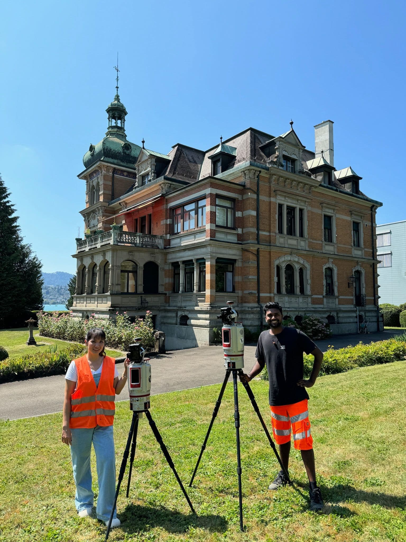 Zwei Vermessungstechniker mit Ausrüstung vor historischem Herrenhaus.