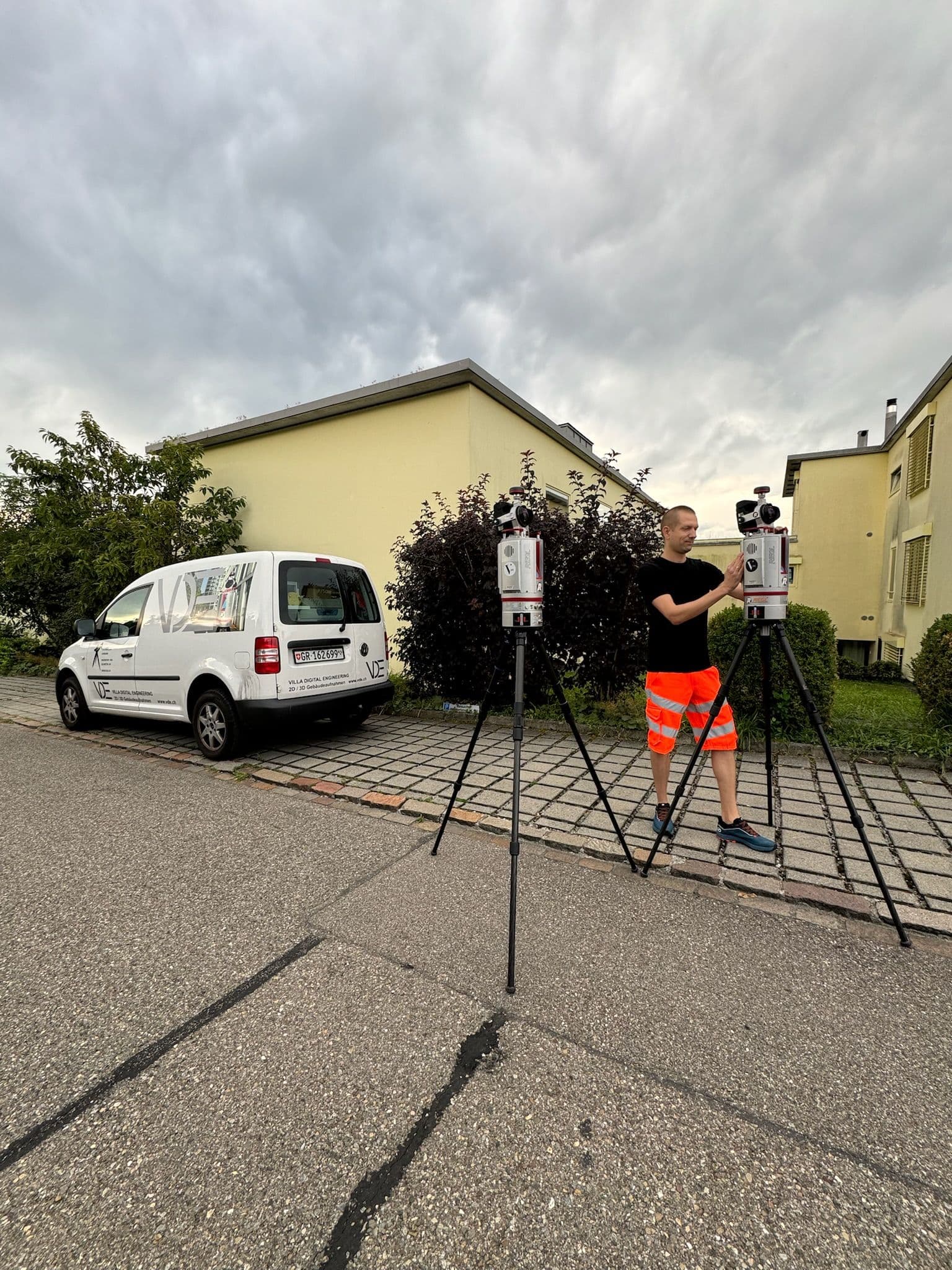 Vermesser mit Theodoliten neben Firmenwagen auf städtischer Straße.