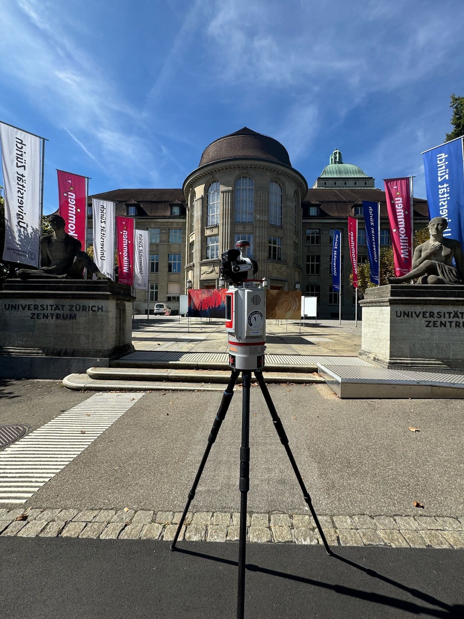 3D-Laserscanner auf Stativ vor dem Haupteingang der Universität Zürich Zentrum, flankiert von Bannern und Steinwänden, im Hintergrund historisches Gebäude unter blauem Himmel.