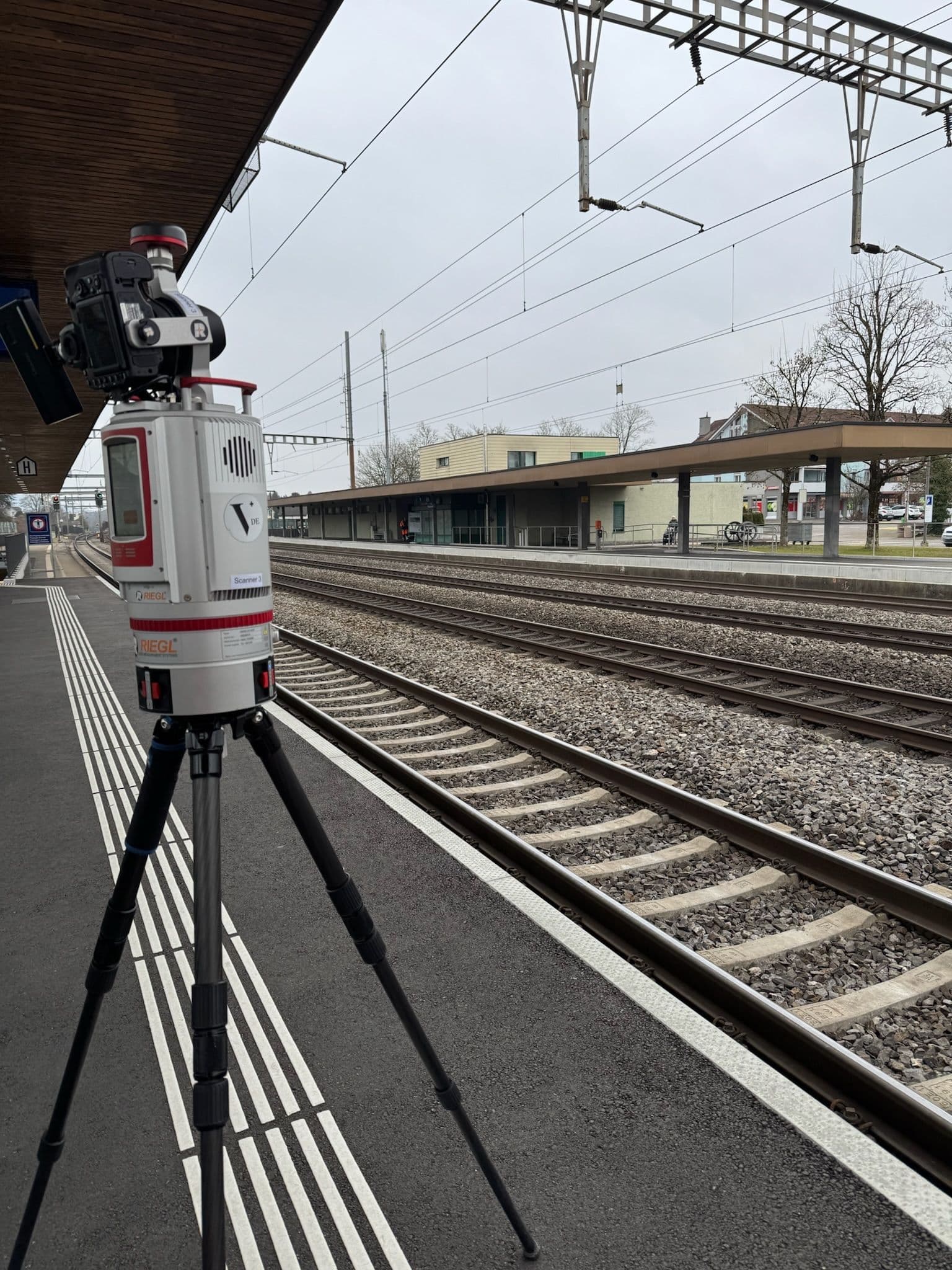 Ein großes Vermessungsgerät steht auf einem Stativ am Bahnsteig eines Bahnhofs, neben Bahngleisen. Der Himmel ist bewölkt und die Umgebung wirkt ruhig.