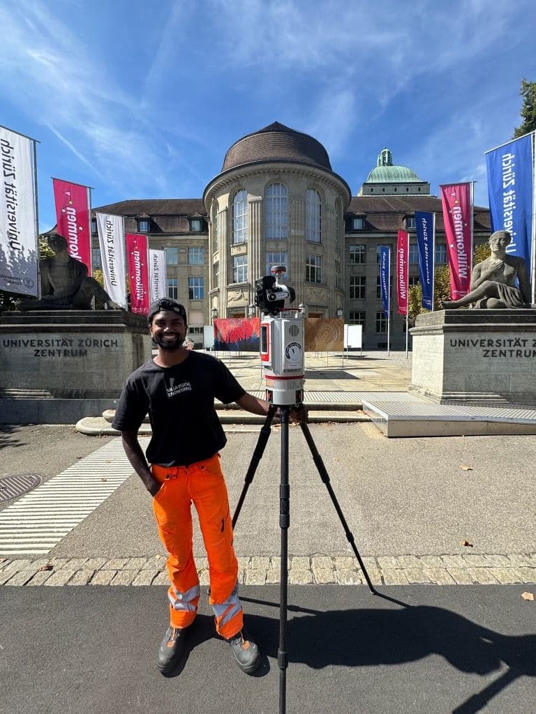 Vermessungsingenieur mit Stativ und 3D-Laserscanner vor der Universität Zürich Zentrum, blauem Himmel und Fahnen; Gebäude und Löwenskulpturen im Hintergrund.