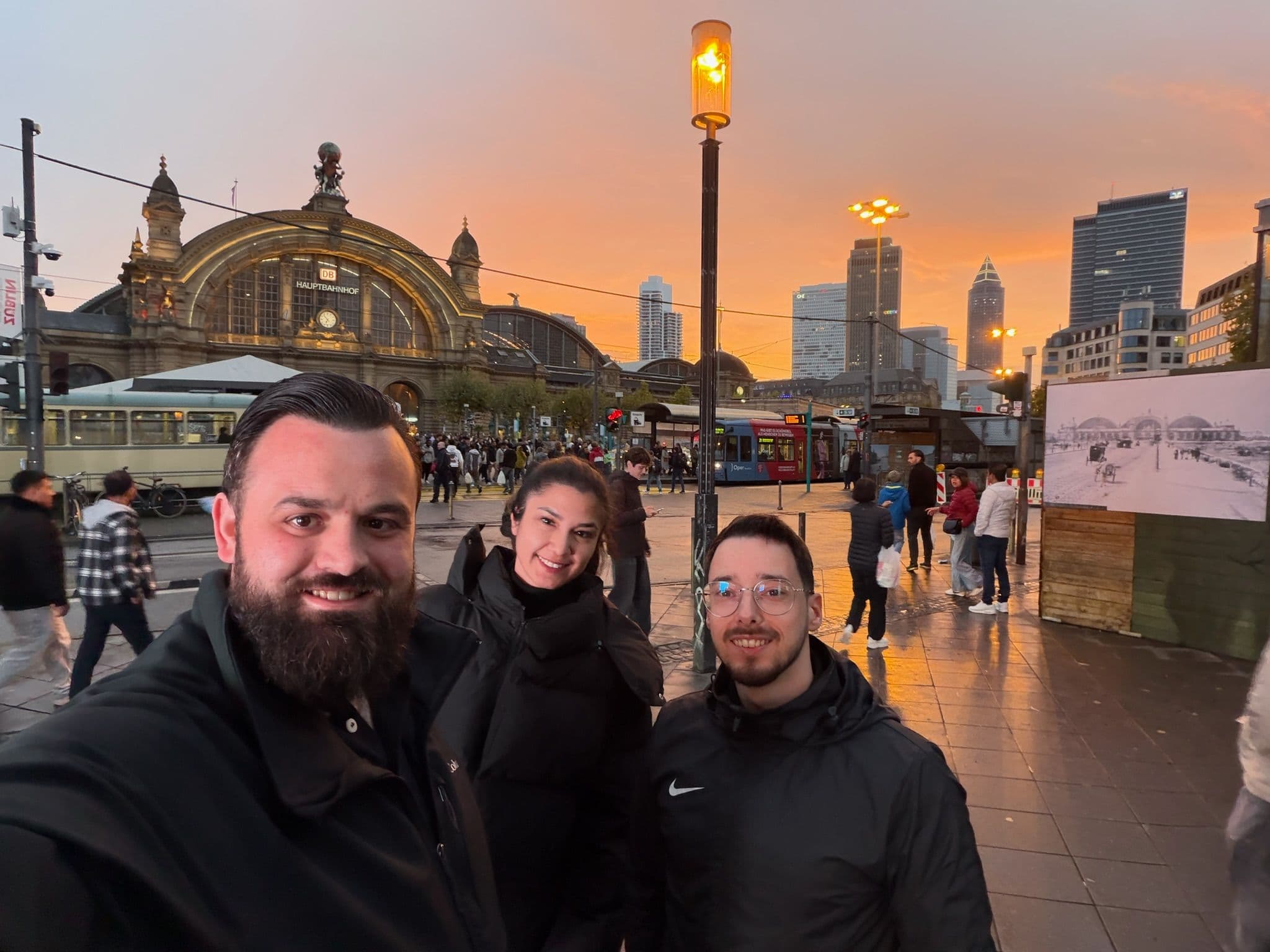 Drei Personen posieren bei Sonnenuntergang vor dem Hauptbahnhof Frankfurt, umgeben von vorbeigehenden Menschen und einer urbanen Kulisse.