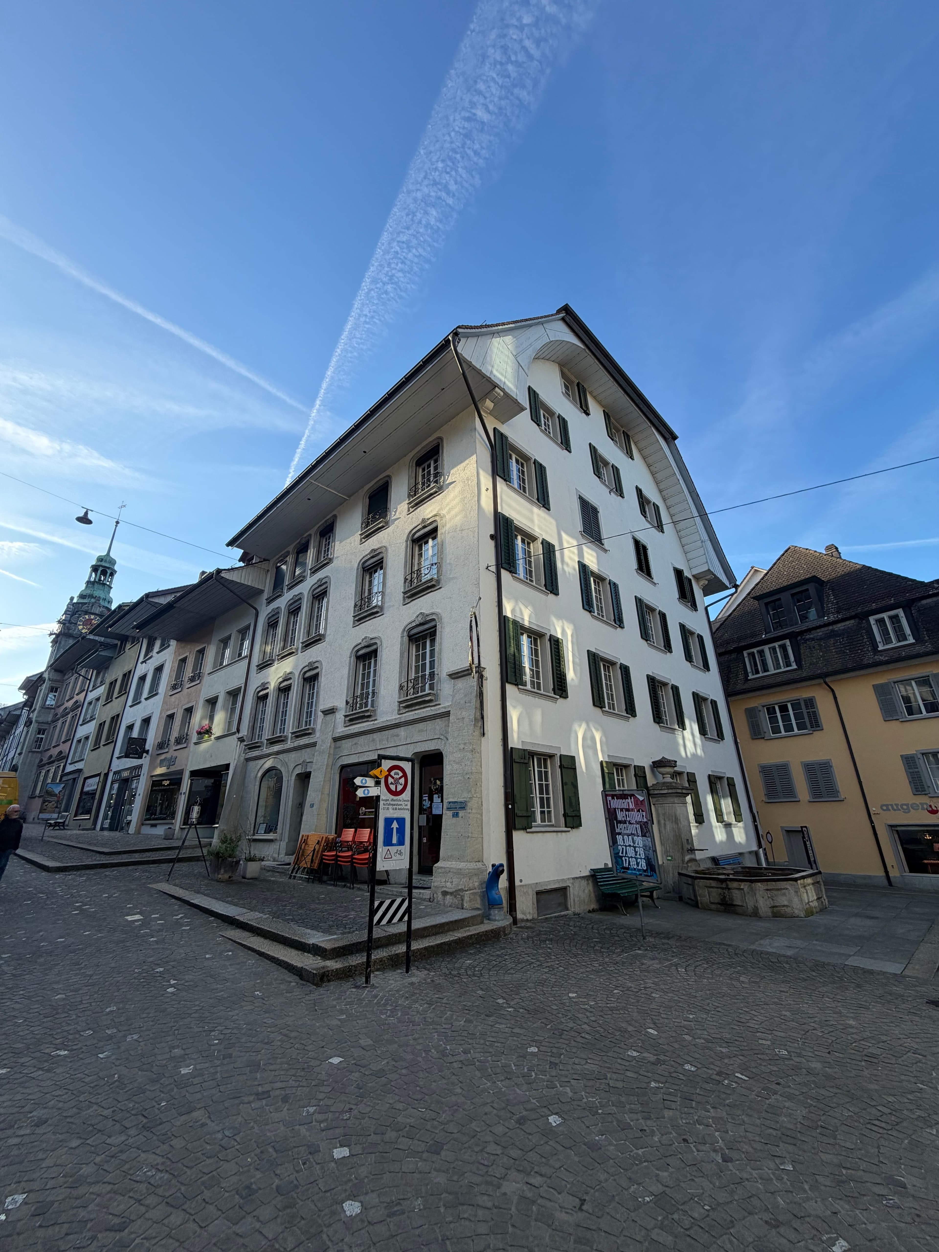 Historisches Altstadtgebäude an gepflastertem Platz mit Straßenschildern und blauem Himmel; perspektivische Weitwinkelaufnahme mit umliegenden Häusern und Kirchturm im Hintergrund.