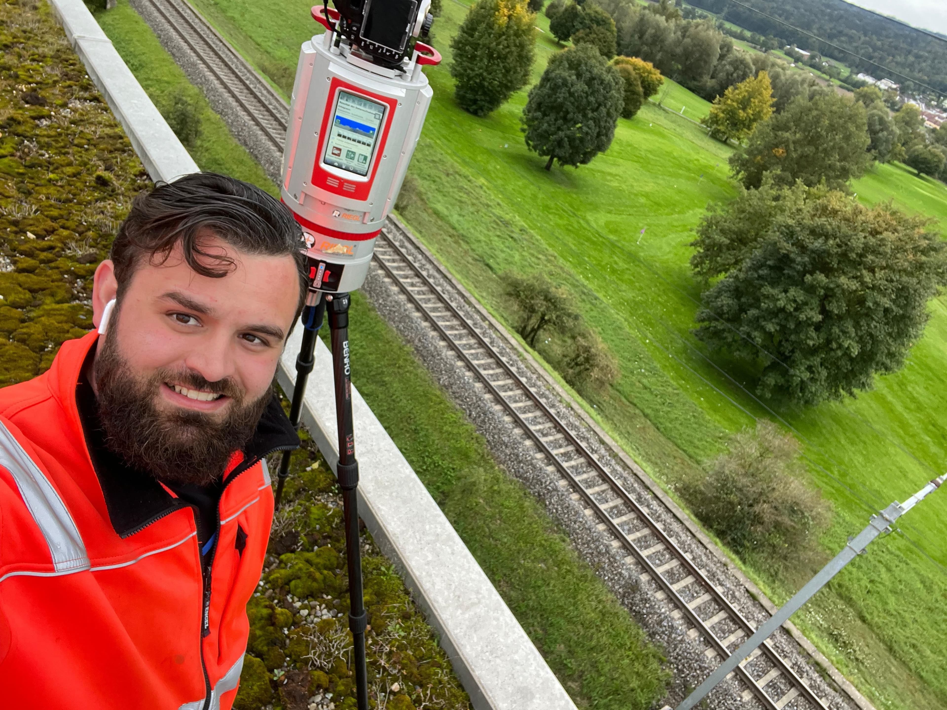 Vermessungstechniker in Warnjacke auf Brücke mit GNSS/Totalstation auf Stativ, Blick auf Bahnstrecke und grüne Landschaft – Gleisvermessung, Bauvermessung, Geodäsie.