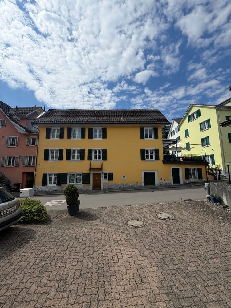 Gelbes Wohnhaus mit schwarzen Fensterläden an einem kleinen Platz, gepflasterter Hof mit Pflanzkübel und geparktem Auto, daneben rosa und grüne Häuser unter blauem Himmel mit Wolken.