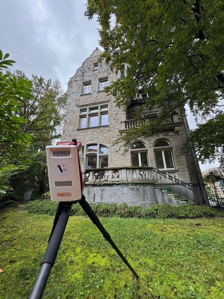 Altes Gebäude mit moderner Vermessungsausrüstung im Vordergrund, umgeben von Bäumen.