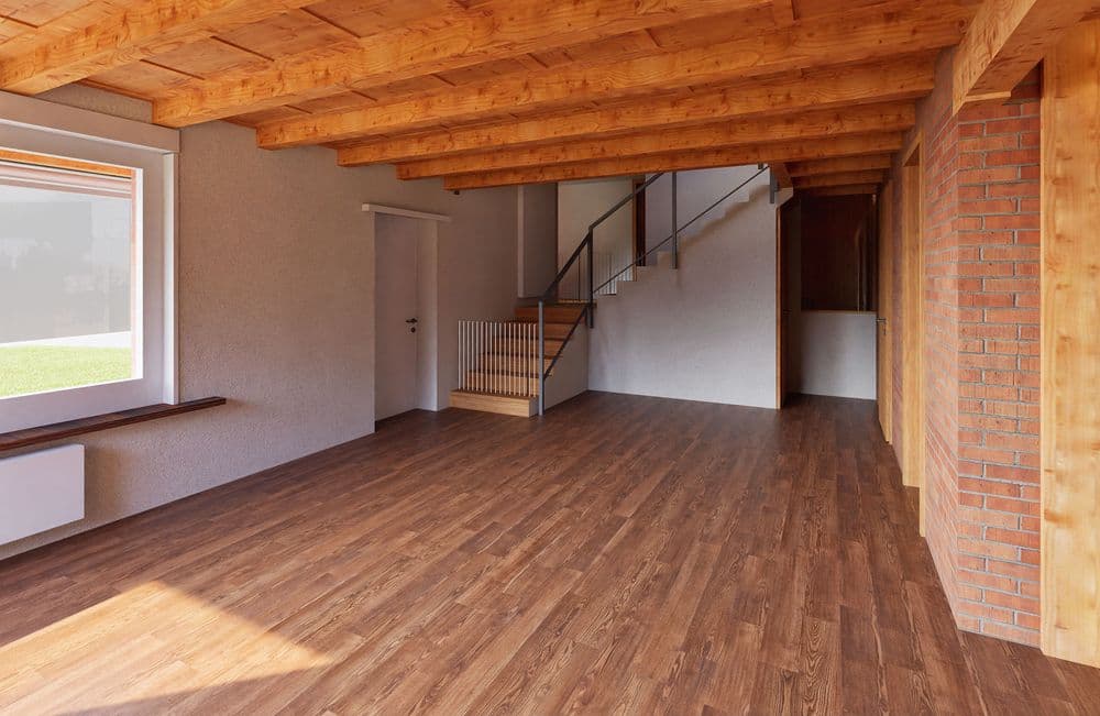 Ein leerer Raum mit Holzdecke und -boden, sichtbaren Ziegelwänden und großer Fensterfront. Moderne Treppe führt nach oben.
