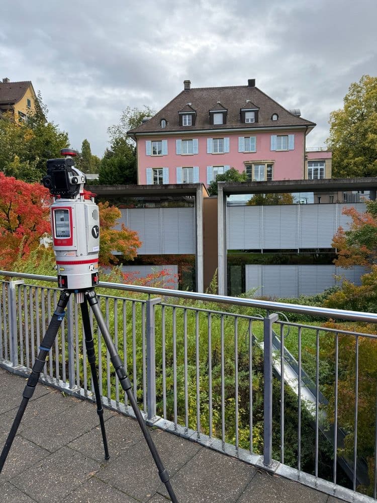 Ein rotes Laserscanner-Gerät auf einem Stativ steht vor einem modernen Gebäude und einem rosa Haus, umgeben von herbstlicher Vegetation.