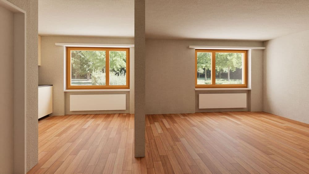 Leerstehender heller Wohnraum mit Holzfußboden, zentraler Stütze, zwei großen Fenstern mit Blick ins Grüne und Heizkörpern darunter, neutral gestrichene Wände.