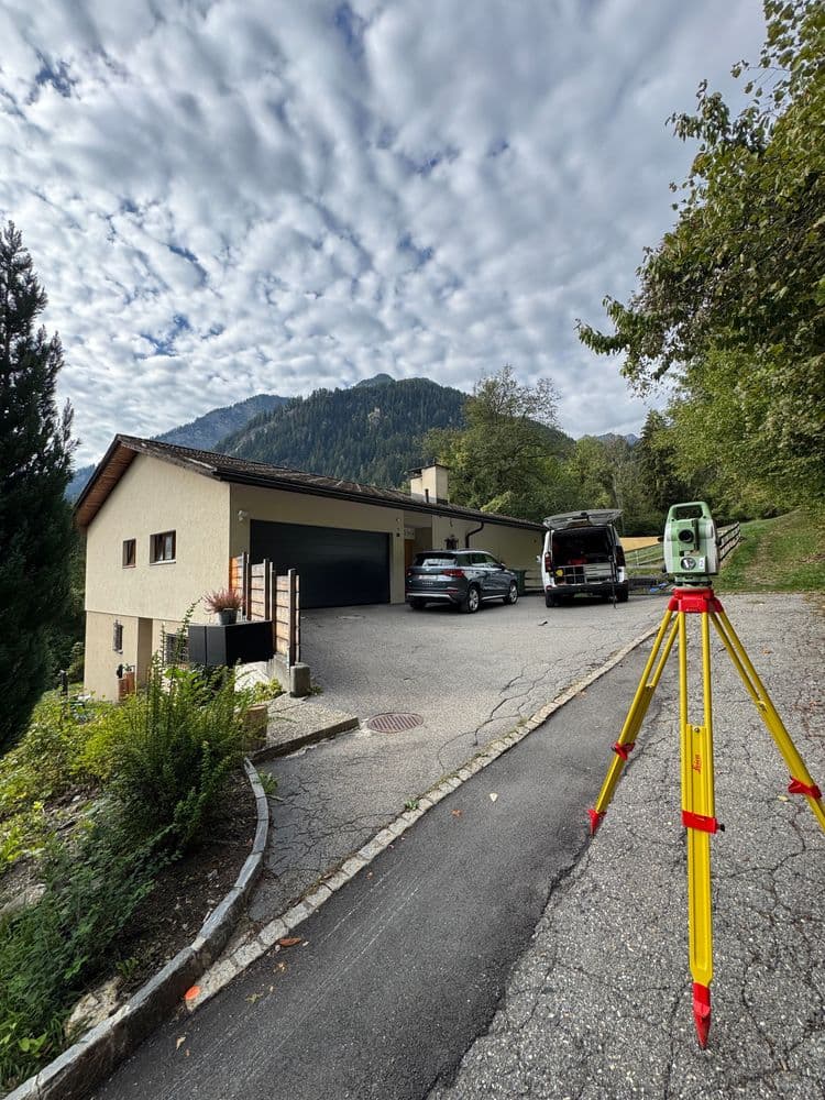 Vermessungsgerät auf Stativ vor Wohnhaus mit Einfahrt und parkenden Autos; im Hintergrund bewaldete Berge und wolkiger Himmel.