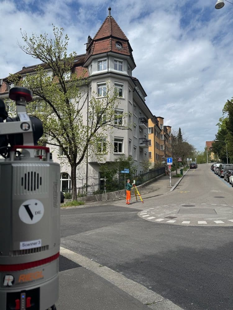 In einem Wohnviertel vermisst ein Arbeiter mit orangener Warnkleidung eine Straße. Im Vordergrund ein Laserscanner, im Hintergrund ein altes Gebäude mit Bäumen.