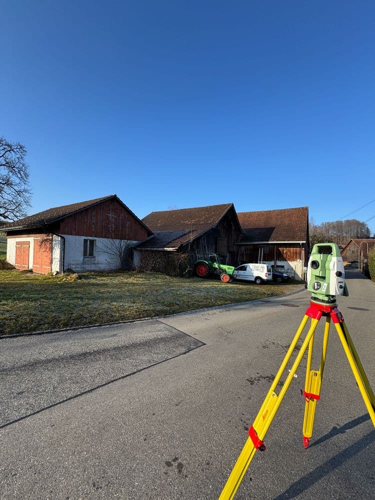 Vermessungsgerät auf Stativ vor Bauernhof mit Scheunen, Traktor und Transporter an ländlicher Straße bei blauem Himmel – geodätische Vermessung auf dem Land.