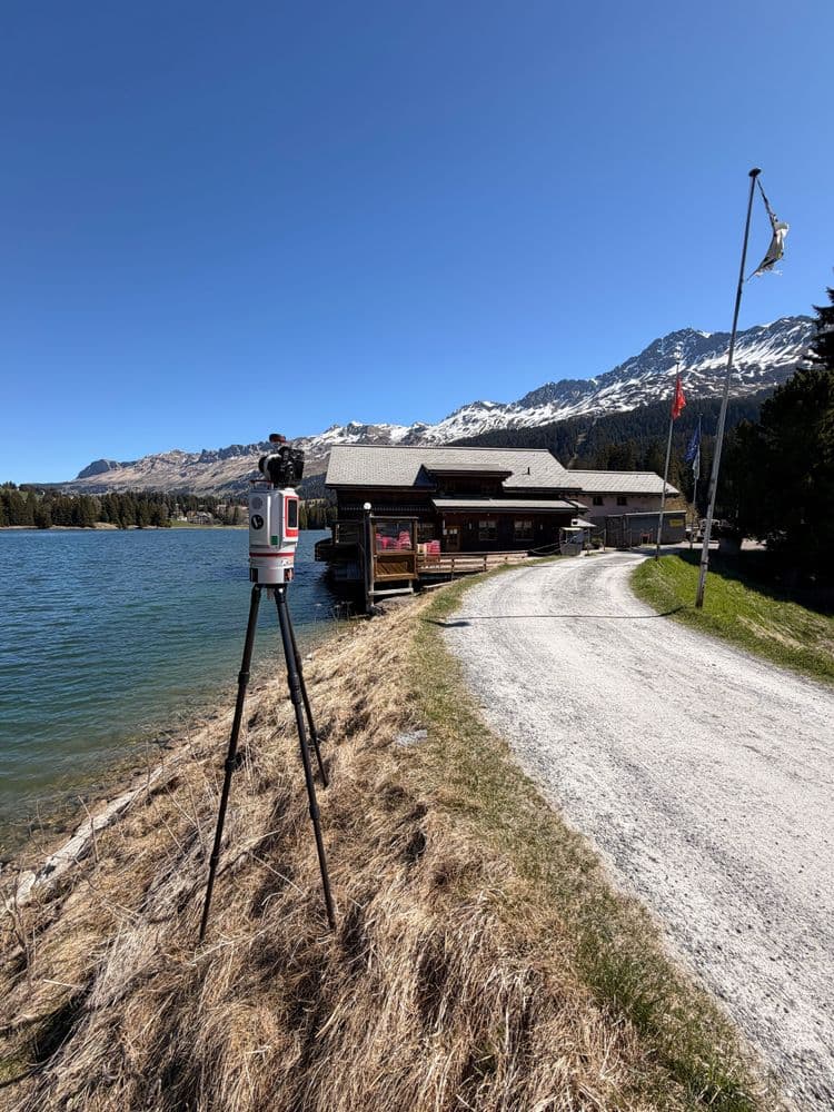 Vermessungsgerät auf Stativ am Ufer eines Bergsees; Schotterweg führt zur Berghütte, dahinter schneebedeckte Alpen unter blauem Himmel.