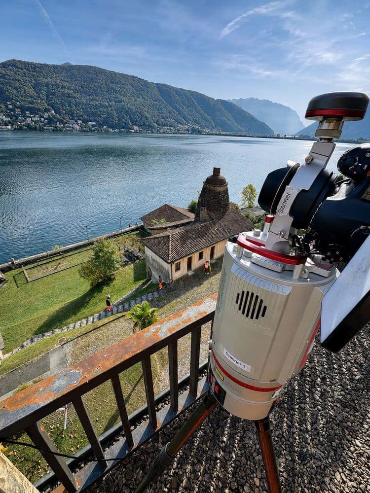 Teleskop auf Stativ auf einer Terrasse mit Blick über einen See und grüne Berge; unten ein historisches Gebäude am Ufer, klarer Himmel und ruhiges Wasser.