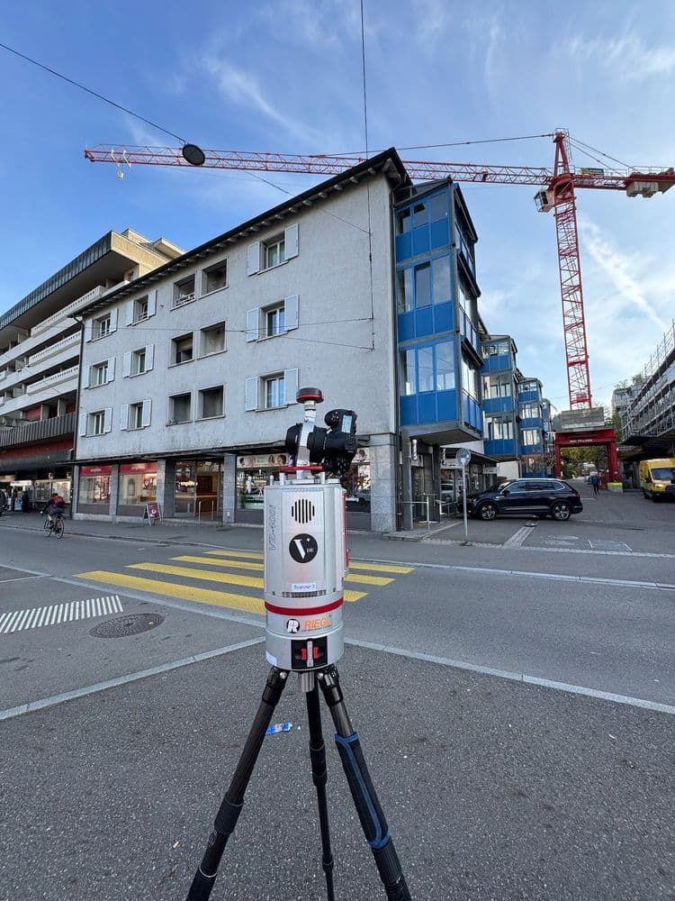 Ein Laserscanner auf einem Stativ misst eine Straßenecke mit mehrstöckigen Gebäuden und Kranauslegern im Hintergrund.