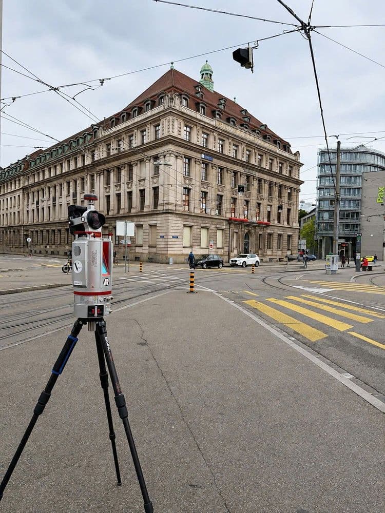 Vermessungsgerät auf Stativ an Straßenkreuzung mit Straßenbahnschienen; im Hintergrund historisches Verwaltungsgebäude und moderne Glasarchitektur in der Stadt bei bewölktem Himmel.