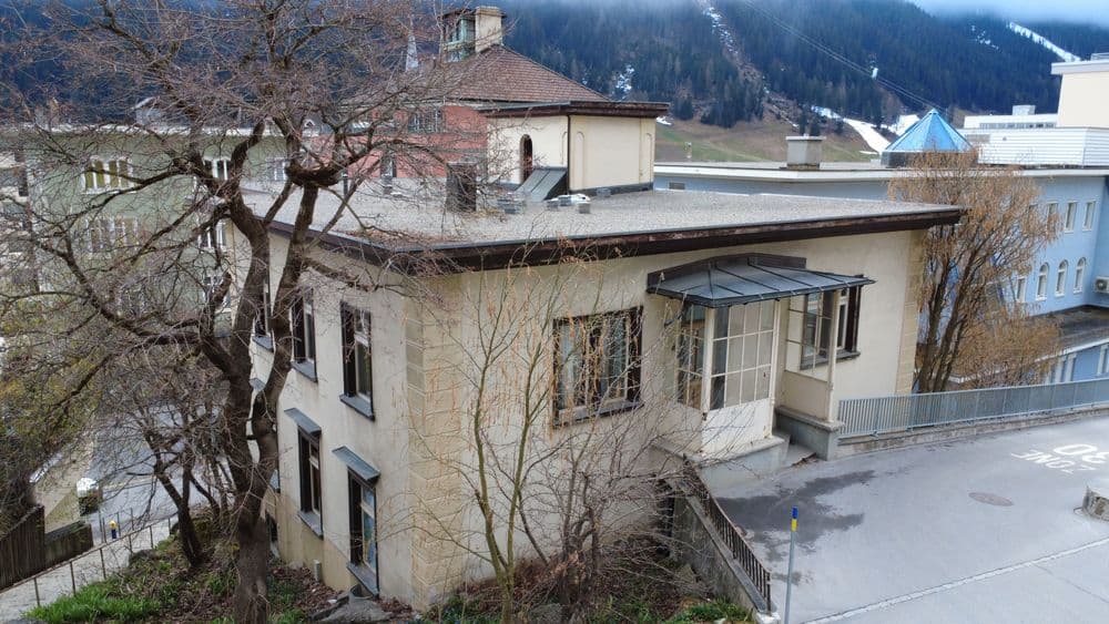 Kleines zweistöckiges Gebäude mit flachem Dach in einer bergigen Umgebung, umgeben von Bäumen und angrenzend an eine Straße.