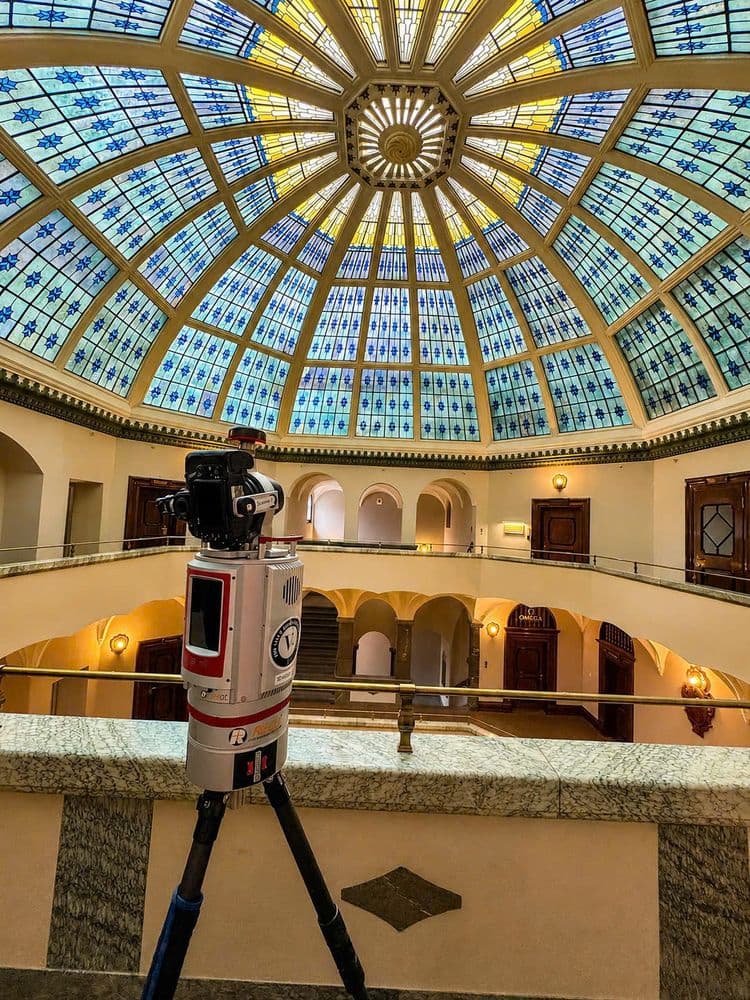 Stativ mit Kamera im Innenhof eines historischen Gebäudes unter großer Glaskuppel mit blauem Buntglas; Rundgalerie, Bögen und Treppenhaus im warmen Licht.