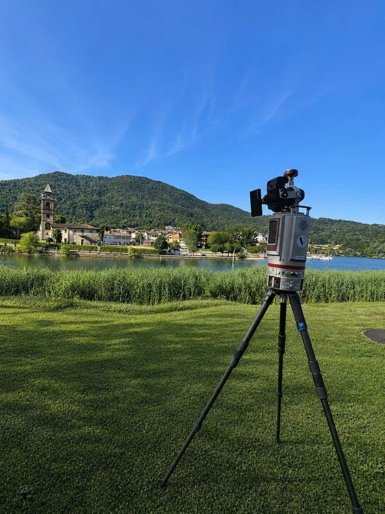 Vermessungsgerät auf Stativ am Seeufer mit Blick auf Dorf und Kirchturm, grüne Wiesen und bewaldete Berge unter blauem Himmel.