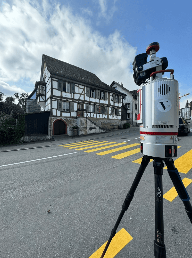 Vermessungsgerät auf Stativ im Vordergrund mit Fachwerkhaus im Hintergrund.