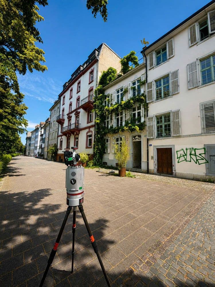 Vermessungsgerät auf Stativ in gepflasterter Straße mit historischen Wohnhäusern, Fensterläden und Begrünung; sonniger Tag, blauer Himmel, Bäume werfen Schatten, Graffiti an Hauswand.