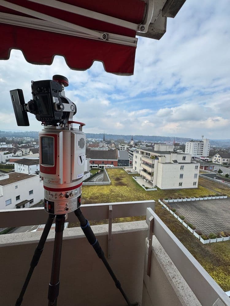 Eine Vermessungsausrüstung steht auf einem Stativ auf einem Balkon und blickt über eine städtische Landschaft mit Wohngebäuden.