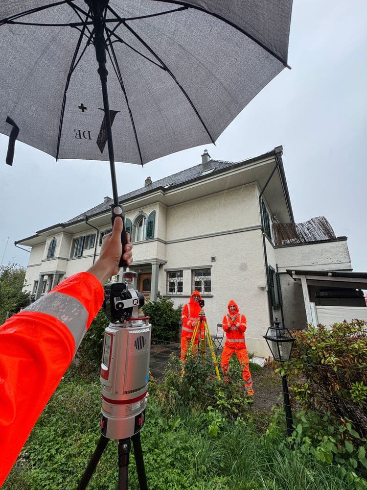 Vermessungsteam in orangefarbener Schutzkleidung vor Wohnhaus im Regen; Hand hält Regenschirm über Messgerät auf Stativ, zwei Vermesser arbeiten im nassen Garten.