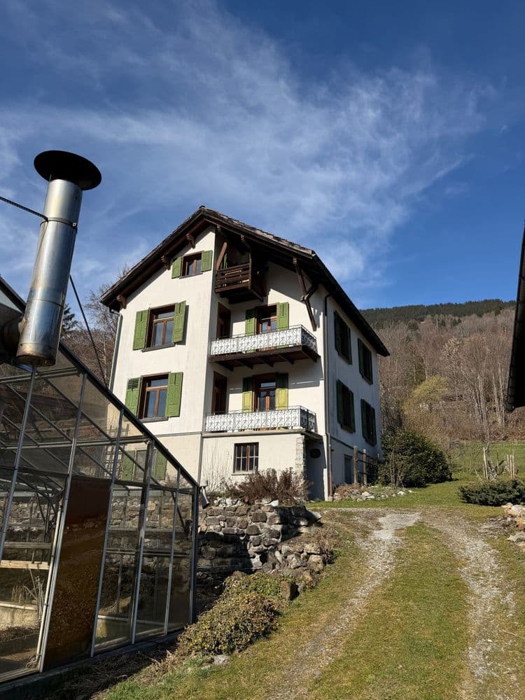 Mehrstöckiges Landhaus mit Balkonen am Hang, daneben Gewächshaus und Schornstein; Schotterweg führt zum Gebäude, blauer Himmel und Waldhügel im Hintergrund.
