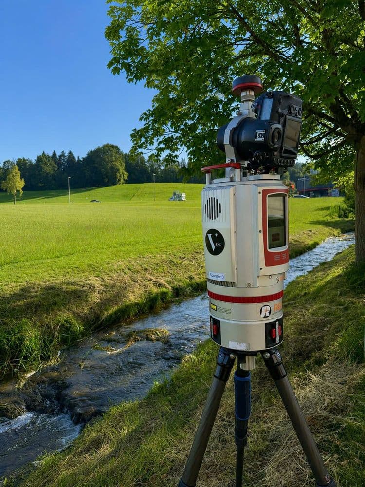 3D-Laserscanner auf Stativ neben Bach am Waldrand, Vermessung in grüner Wiesenlandschaft bei Sonnenschein und blauem Himmel.