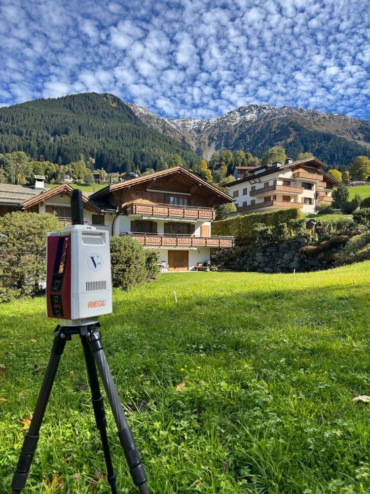 3D-Laserscanner auf Stativ auf grüner Wiese vor alpinen Chalets; dahinter bewaldete Berge und Gipfel unter wolkigem Himmel – Vermessung und Bauaufnahme in den Alpen.