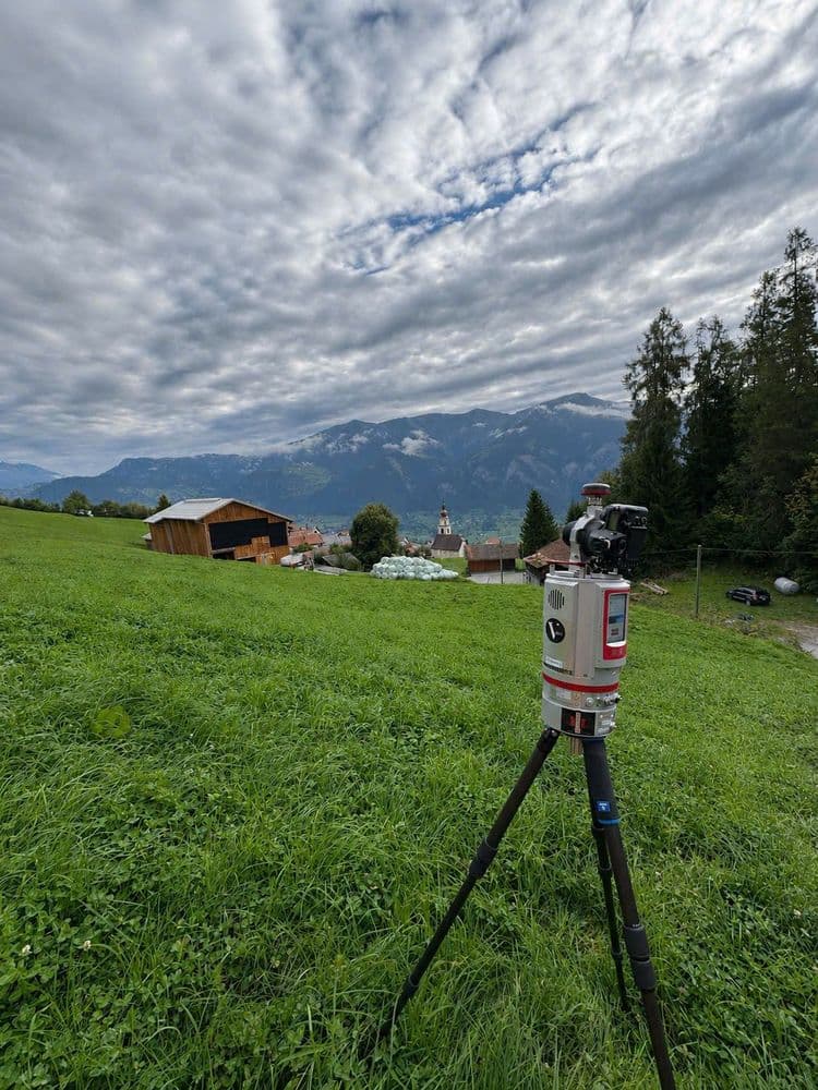 Vermessungsgerät auf Stativ in grüner Alpenwiese, dahinter Bauernhof und Dorf mit Kirchturm vor Bergpanorama unter dramatischem Wolkenhimmel.