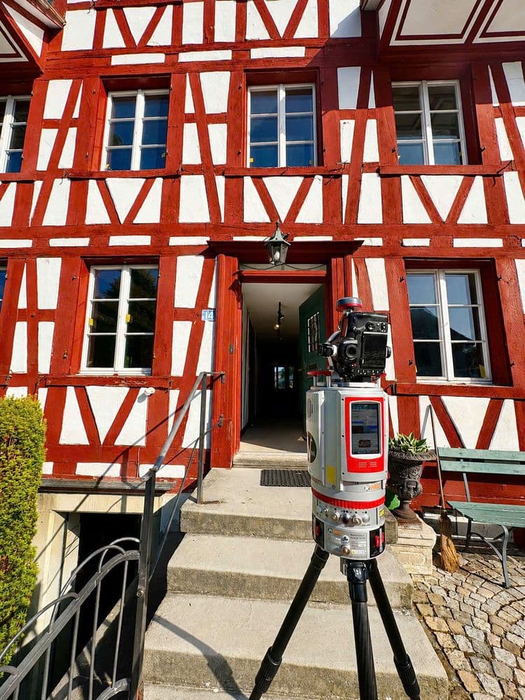Rotes Fachwerkhaus mit weißem Putz und Eingangstreppe; davor steht ein 3D-Laserscanner auf Stativ zur Gebäudevermessung.