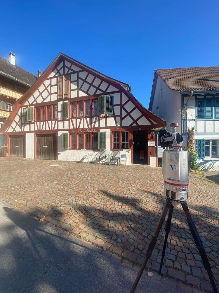 Fachwerkhaus mit roten und weißen Details, daneben ein modernes Gerät auf Stativ, gepflasterter Platz im Vordergrund.