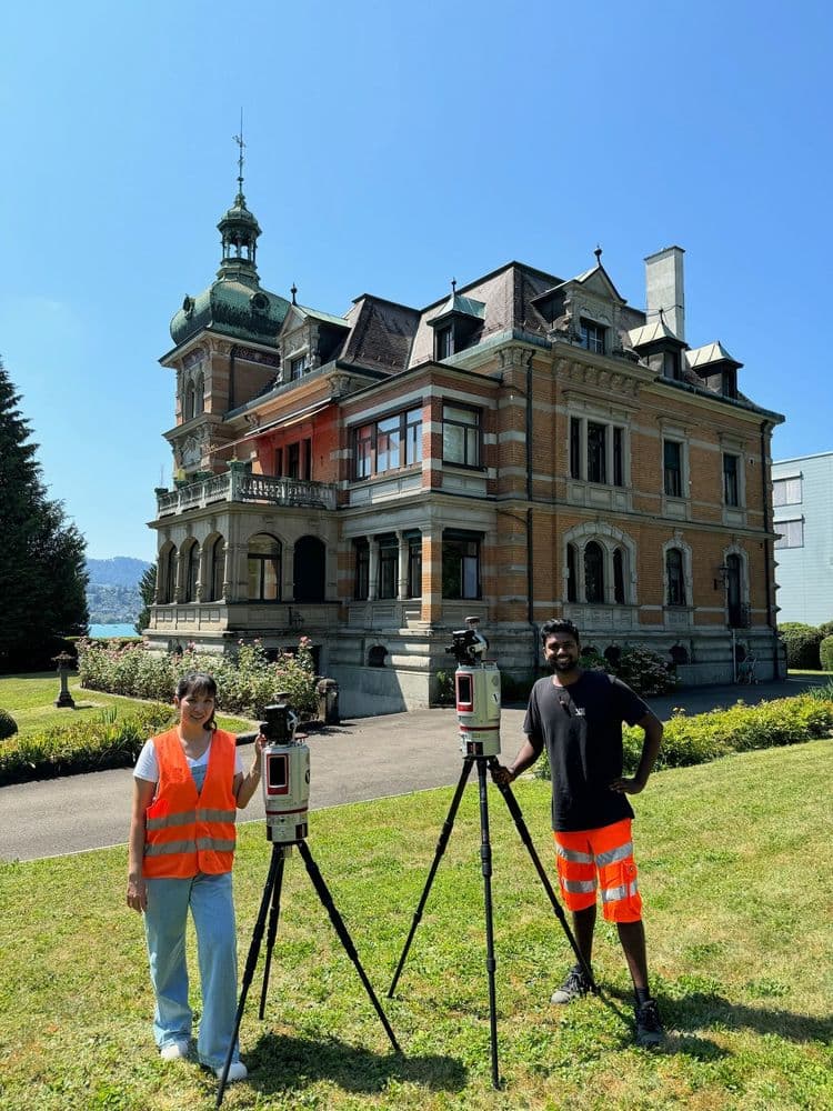 Zwei Vermessungstechniker mit Ausrüstung vor historischem Herrenhaus.