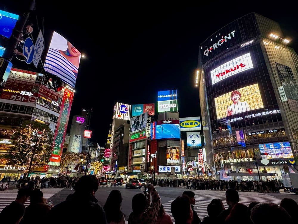 Der menschenbefüllte Shibuya-Kreuzung in Tokio bei Nacht, umgeben von hell erleuchteten Werbetafeln und hohen Gebäuden.