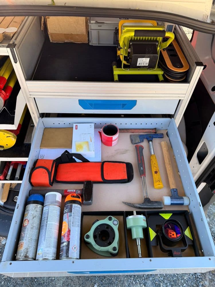 Offene Werkzeugschublade in einem Servicefahrzeug mit Spraydosen, Hammer, Meißel, Maßband, Markierungsband, Halterungen und Akku-Arbeitslampe – ordentlich sortierte Werkstattausrüstung im Transporter.