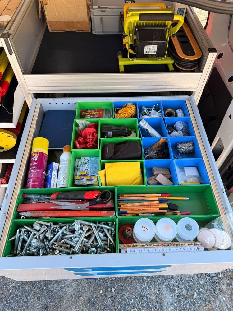 Offene Werkzeugschublade im Werkstattwagen mit sortierten Schrauben, Bits, Kleinteilen, Klebeband, Stiften, Zangen und Messwerkzeugen in farbigen Fächern.