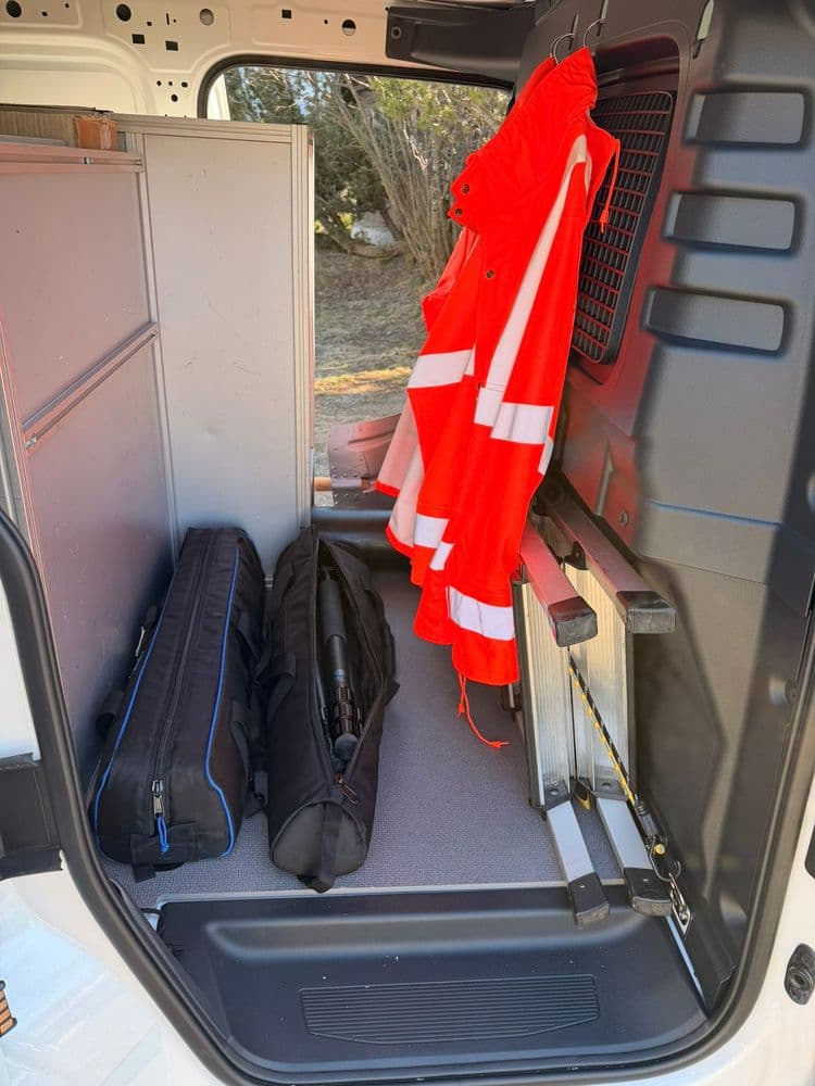 Laderaum eines Transporters mit hängender roter Warnjacke, Werkzeugkoffer und Taschen; seitliche Schränke und Regalsystem für Handwerker-Ausrüstung.