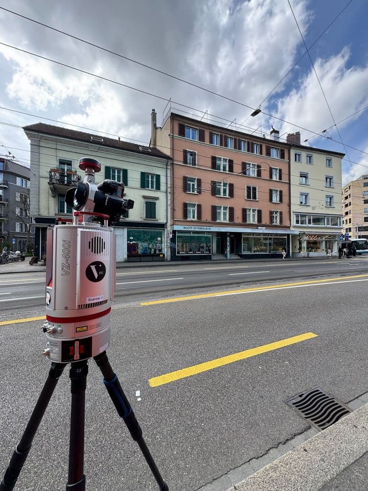 Tragbares Lasermessgerät auf Stativ vor einer mehrstöckigen Häuserzeile in der Stadt, mit Wolken am Himmel.
