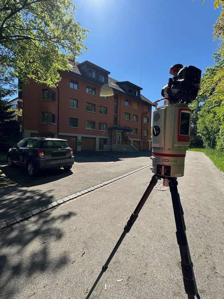In einem von Bäumen gesäumten Wohngebiet steht ein rotes Gebäude. Im Vordergrund befindet sich ein Vermessungsgerät auf einem Stativ, während ein Auto in der Nähe des Hauses geparkt ist.