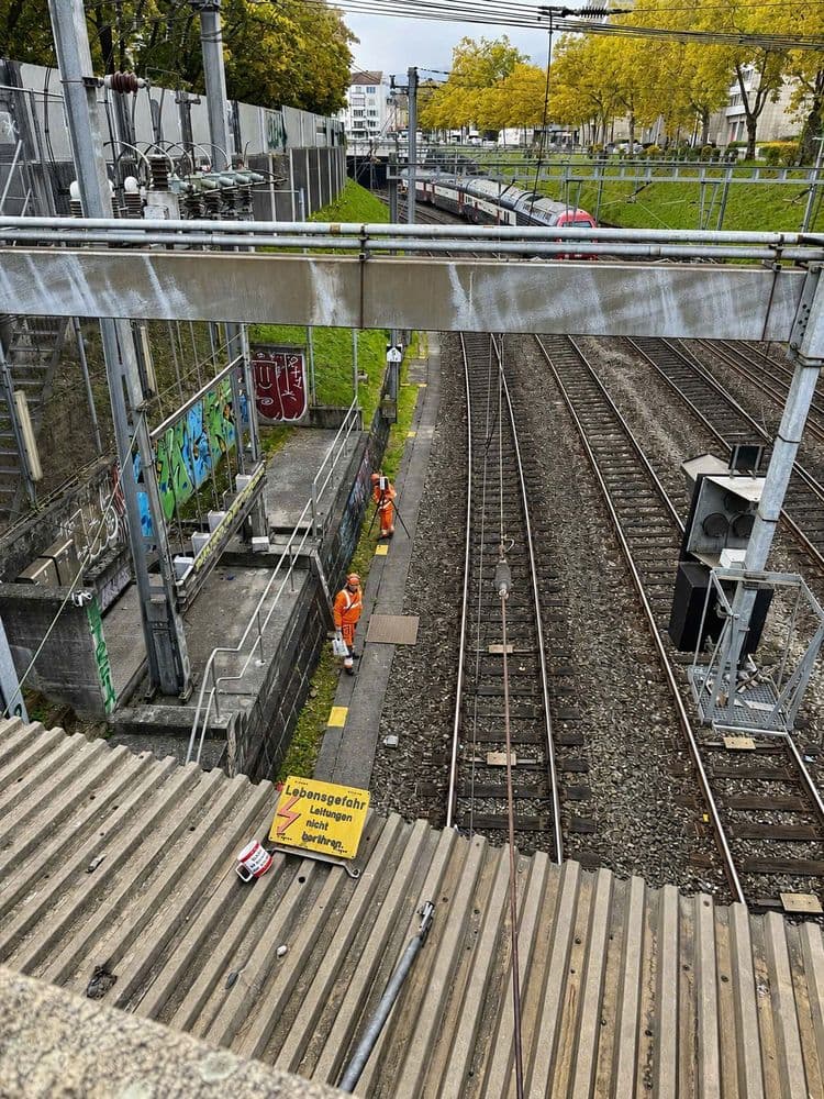 Blick von einer Brücke auf mehrere Bahngeleise neben einer Baustelle; zwei Arbeiter in orange arbeiten am Gleis, umgeben von Oberleitungen, Signaltechnik, Graffitiwand und herbstlichen Bäumen.