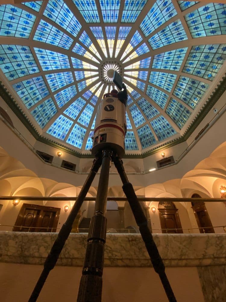 Kuppel mit blauem Glasfenster, darunter ein Fernglas auf einem Stativ, umgeben von einer Marmorbalustrade in einem historischen Gebäude.