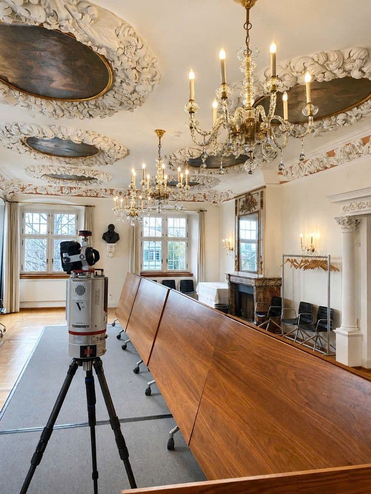 Historischer Konferenzraum mit Stuckdecke und Kronleuchtern, langem Holztisch, Stühlen, Kamin und großen Fenstern; im Vordergrund steht ein Vermessungsgerät auf Stativ.