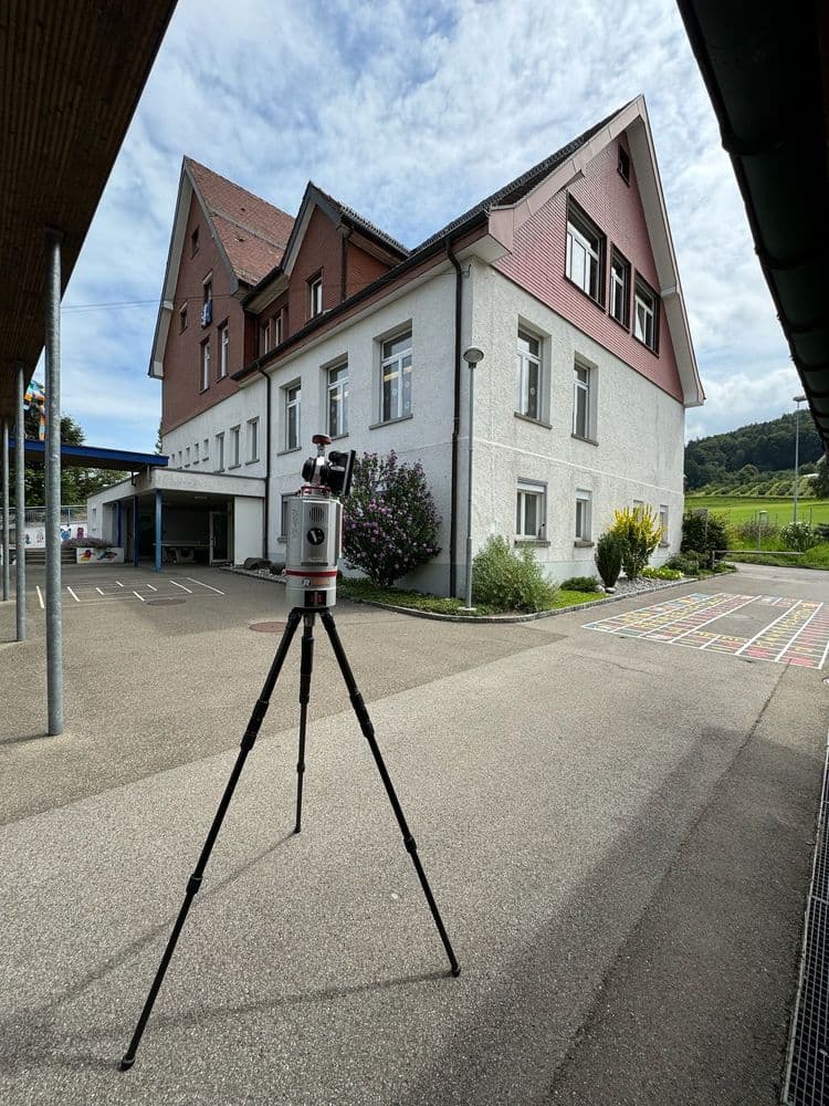 Vermessungsgerät auf Stativ vor einem Schulgebäude mit rotem Dach und blauem Himmel.