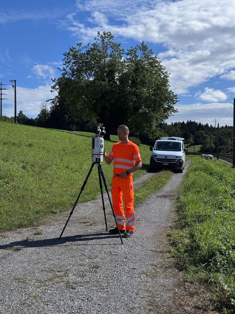 Vermessungstechniker in orangefarbener Warnkleidung bedient Messgerät auf Stativ an Feldweg; im Hintergrund weißer Transporter, grüne Wiesen und blauer Himmel mit Wolken.
