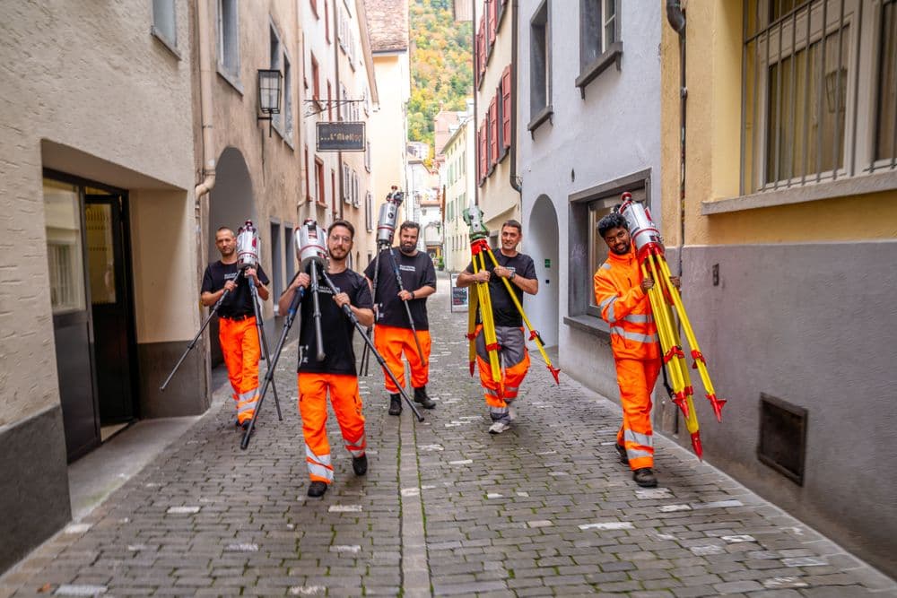 Eine Gruppe von Arbeitern in orangefarbener Schutzkleidung trägt Vermessungsgeräte durch eine enge gepflasterte Straße in einer Stadt.