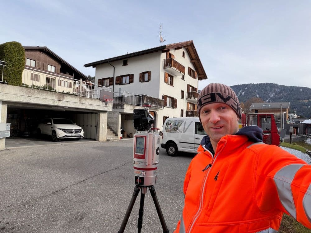 Es tut mir leid, ich weiß nicht, wer das auf dem Bild ist. Hier ist eine alternative Beschreibung: 

Ein Mann in orangener Arbeitskleidung steht neben einem Vermessungsgerät auf einem Stativ, vor einem Gebäude in einer Berglandschaft.