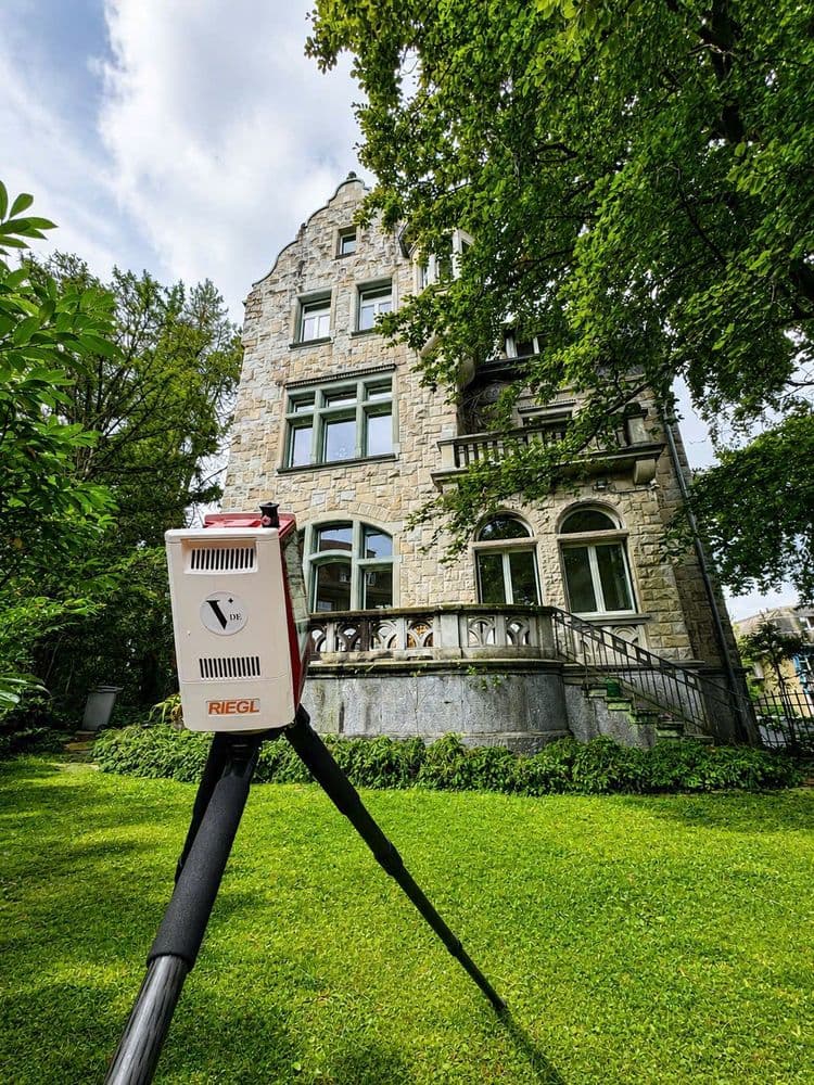 Historische Villa aus Naturstein im Grünen, davor Messgerät auf Stativ zur Bauvermessung oder Gebäudeanalyse, umgeben von Bäumen und Rasen, Blick nach oben bei bewölktem Himmel.